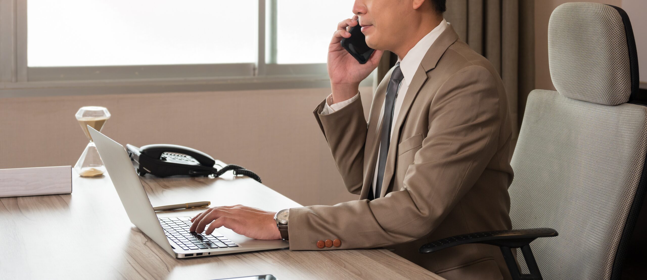 Asian businessman in formalwear talking on smart phone and analyzing  financial development strategy data with laptop computer while sitting on desk in modern private office